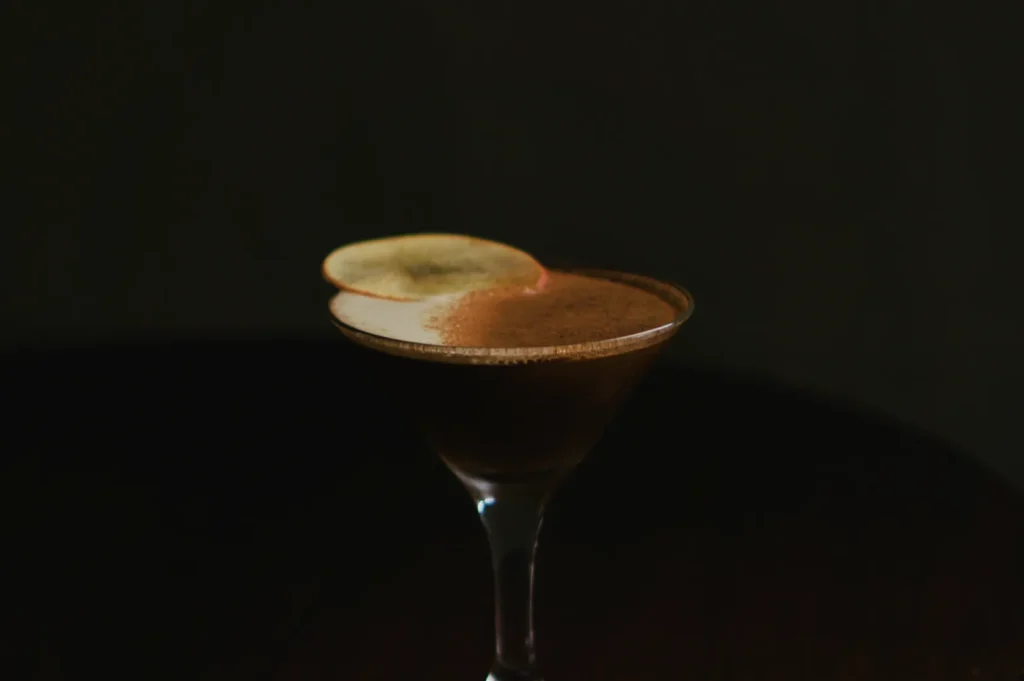 A dark cocktail in a martini glass with a frothy top and a thin lemon slice garnish, set against a moody dark background on a wooden surface.