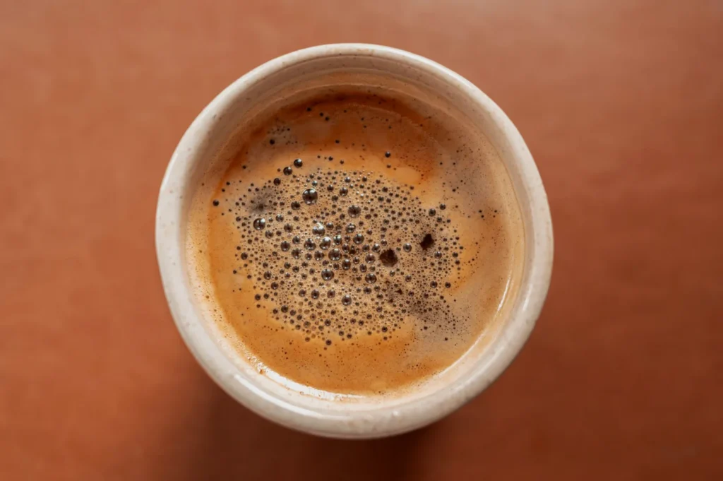 “Top view of a ceramic cup filled with freshly brewed coffee, featuring a rich crema and bubbles on the surface, set against a warm brown background.”