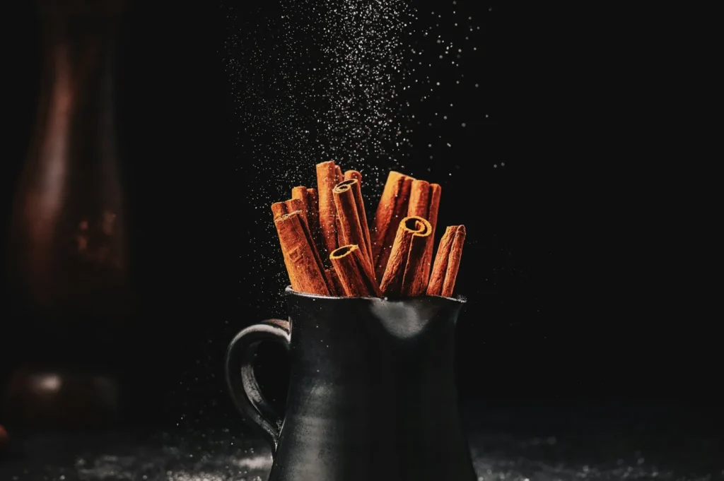 Cinnamon sticks in a dark, ceramic jug with sugar dusting them, set against a black background. The scene evokes a warm, rustic feeling.