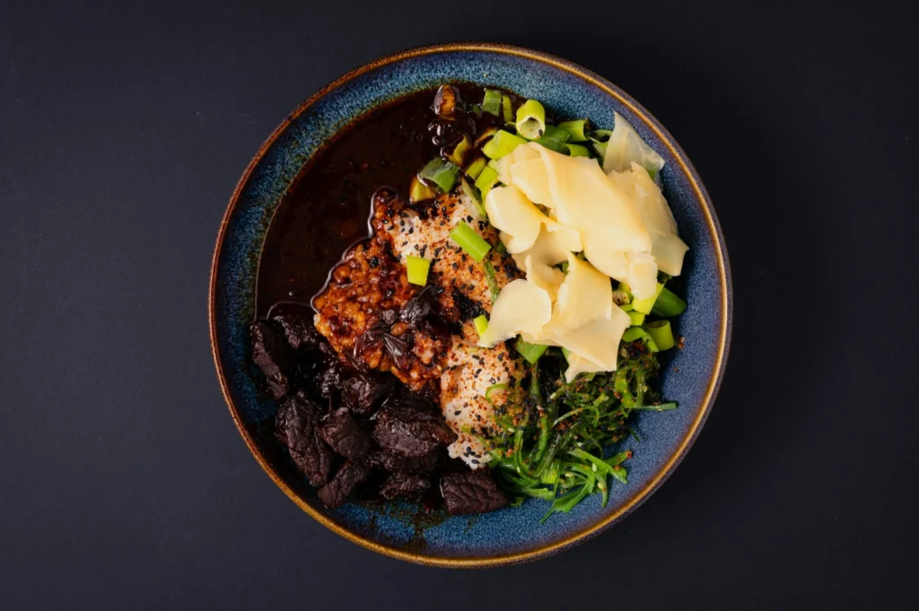 “A vibrant ceramic bowl on a dark background filled with slices of beef, ginger, green onions, seaweed, and a spicy red sauce, creating a rich, savory presentation.”