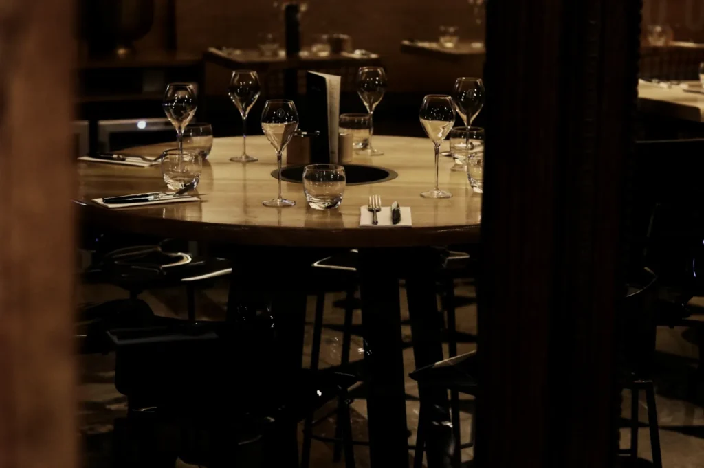 A dimly lit restaurant table set with empty wine glasses and small water glasses, surrounded by dark chairs. The mood is elegant and intimate.
