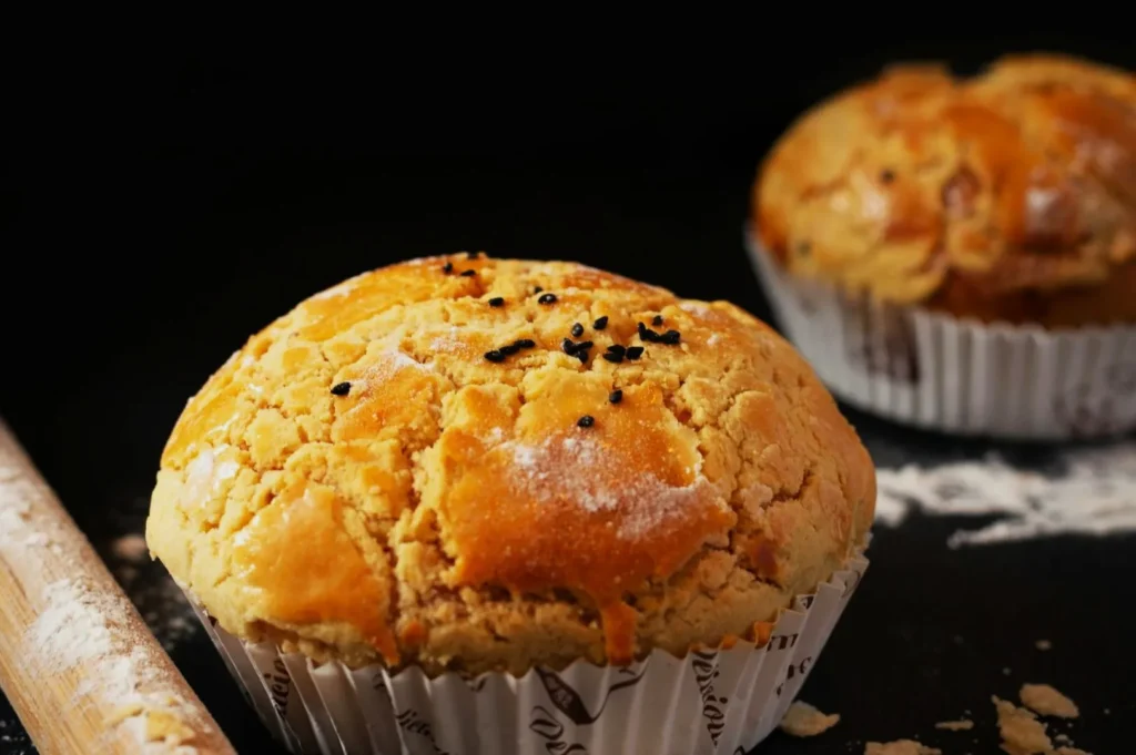 “Close-up of a golden brown muffin with a cracked crust, sprinkled with black sesame seeds, on a dark background. Another muffin and flour dust are in the background.”