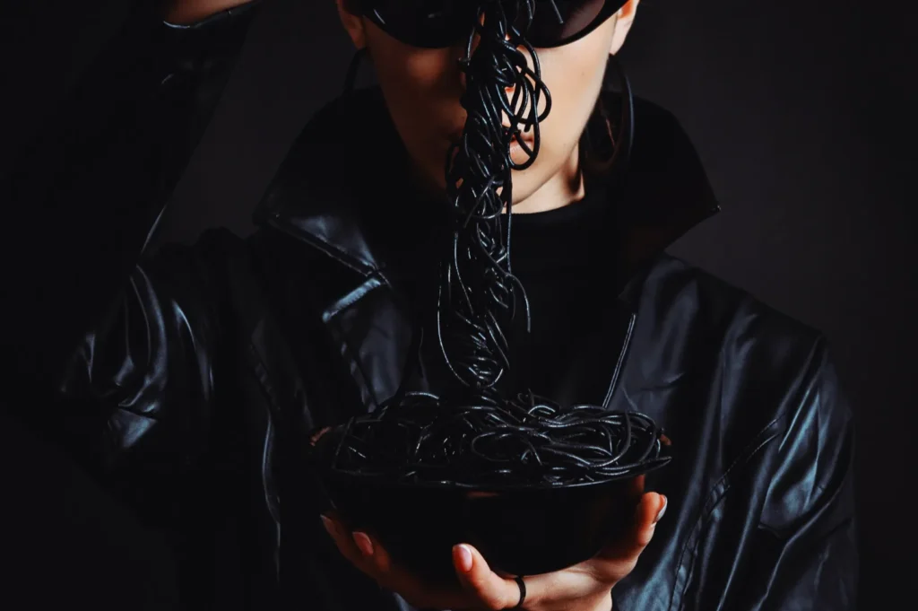 “A person in dark sunglasses and a leather jacket holds a bowl of black spaghetti, lifting a forkful. The mood is edgy and mysterious.”
