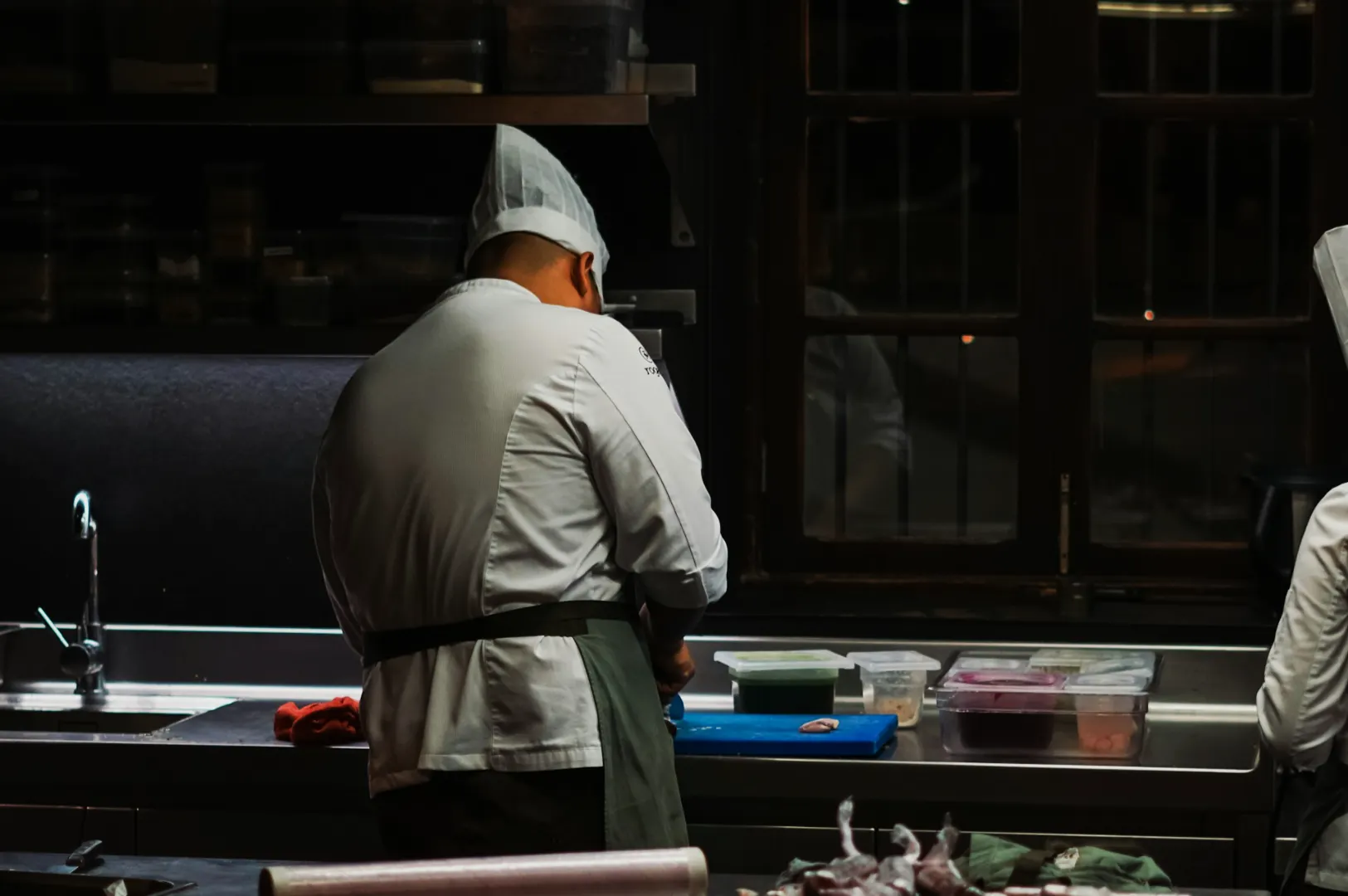Chef in a white uniform and hat working in a dimly lit kitchen, focused on a prep station with containers and a blue cutting board, creating a calm ambiance.