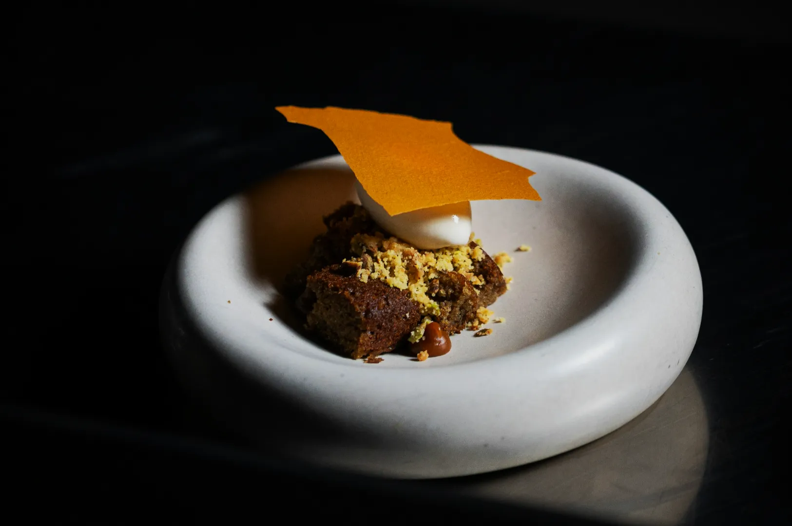A gourmet dessert on a white, shallow dish, featuring crumbled cake, a scoop of ice cream, and an orange wafer topping, set on a dark surface.