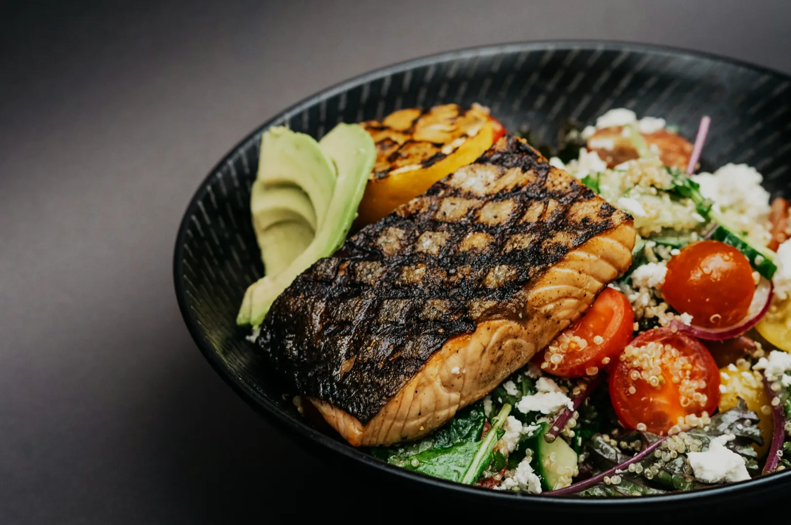 Grilled salmon with char marks rests on a bed of quinoa salad with cherry tomatoes, spinach, grilled vegetables, avocado slices, and crumbled feta.