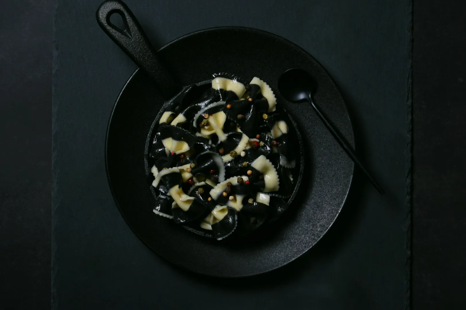 Black and white bowtie pasta with peppercorn garnish in a black pan. A black spoon is placed beside, conveying a modern, minimalist tone.