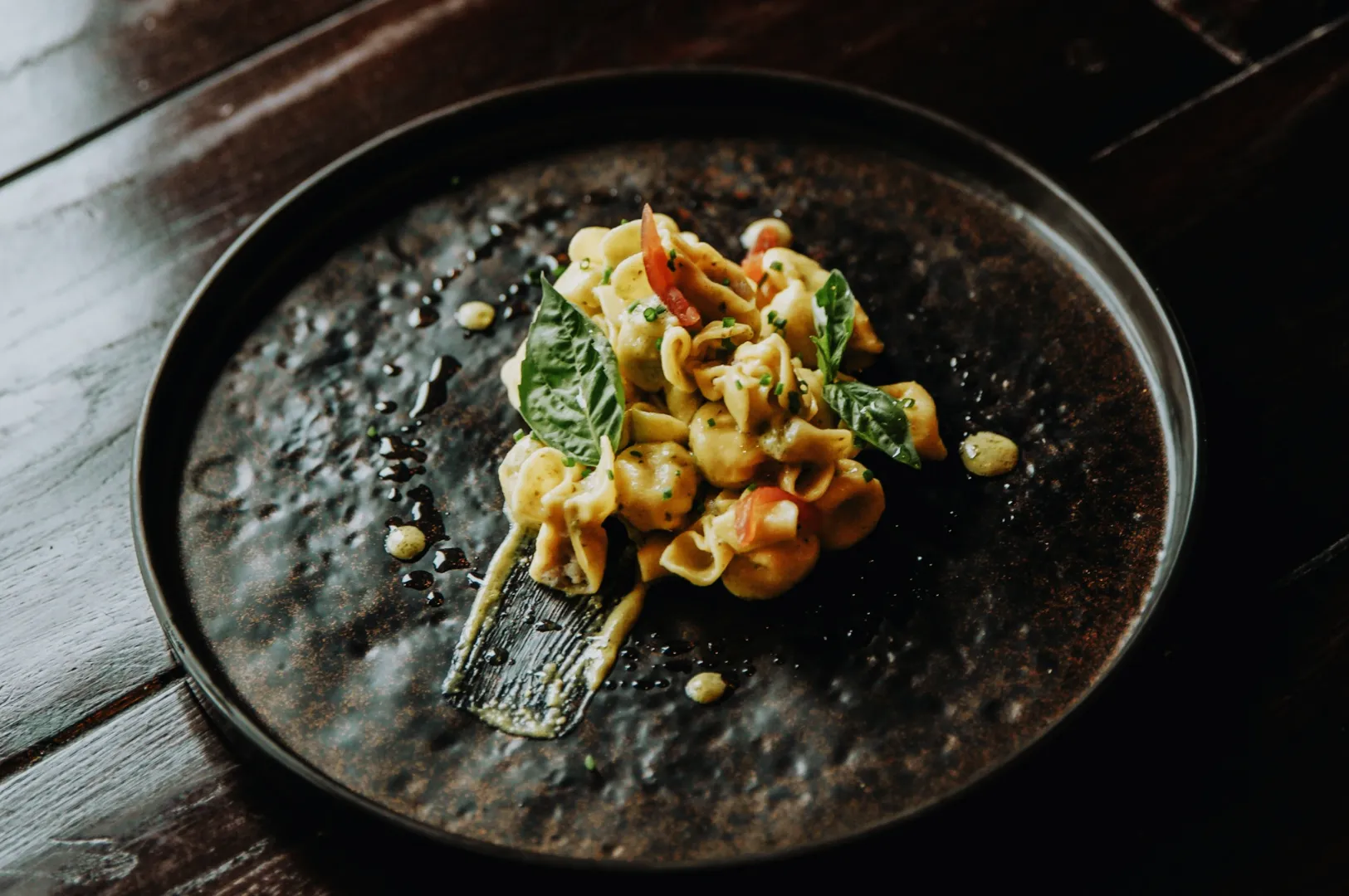 A dark ceramic plate with tortellini in creamy sauce, garnished with fresh basil and diced tomatoes on a rustic wooden table. Elegant and appetizing.