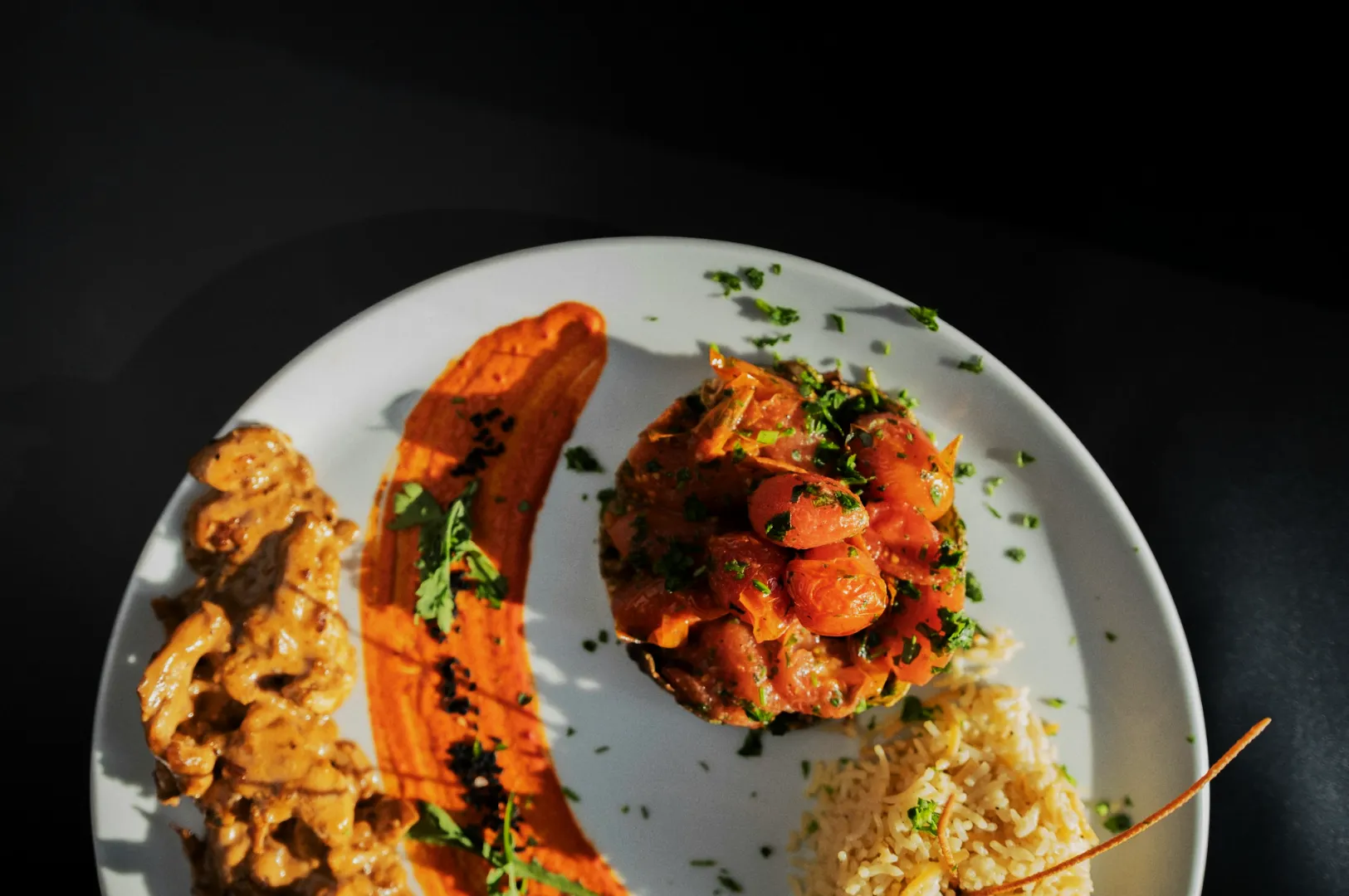 A white plate with a visually appealing dish: creamy beef strips, vibrant orange sauce, fresh tomato salad, and seasoned rice, garnished with herbs.