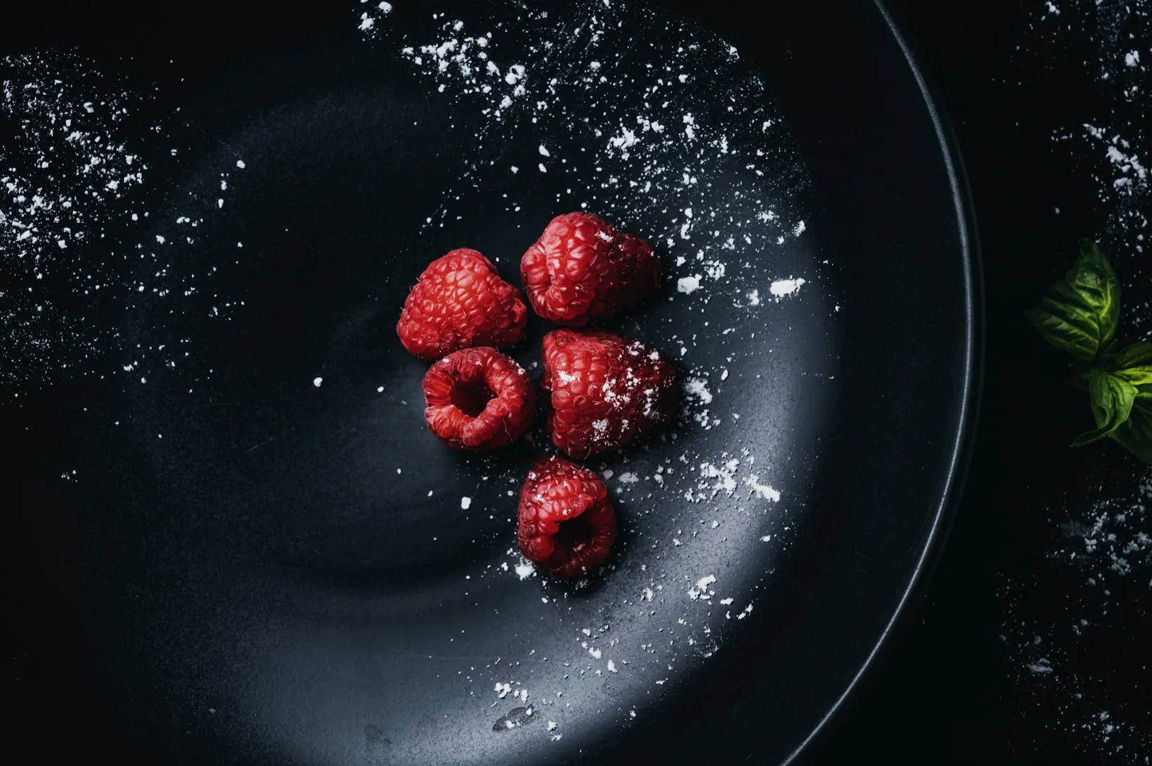 Five raspberries dusted with powdered sugar on a dark plate, creating a striking contrast. A few green leaves peek in from the side, adding a fresh touch.