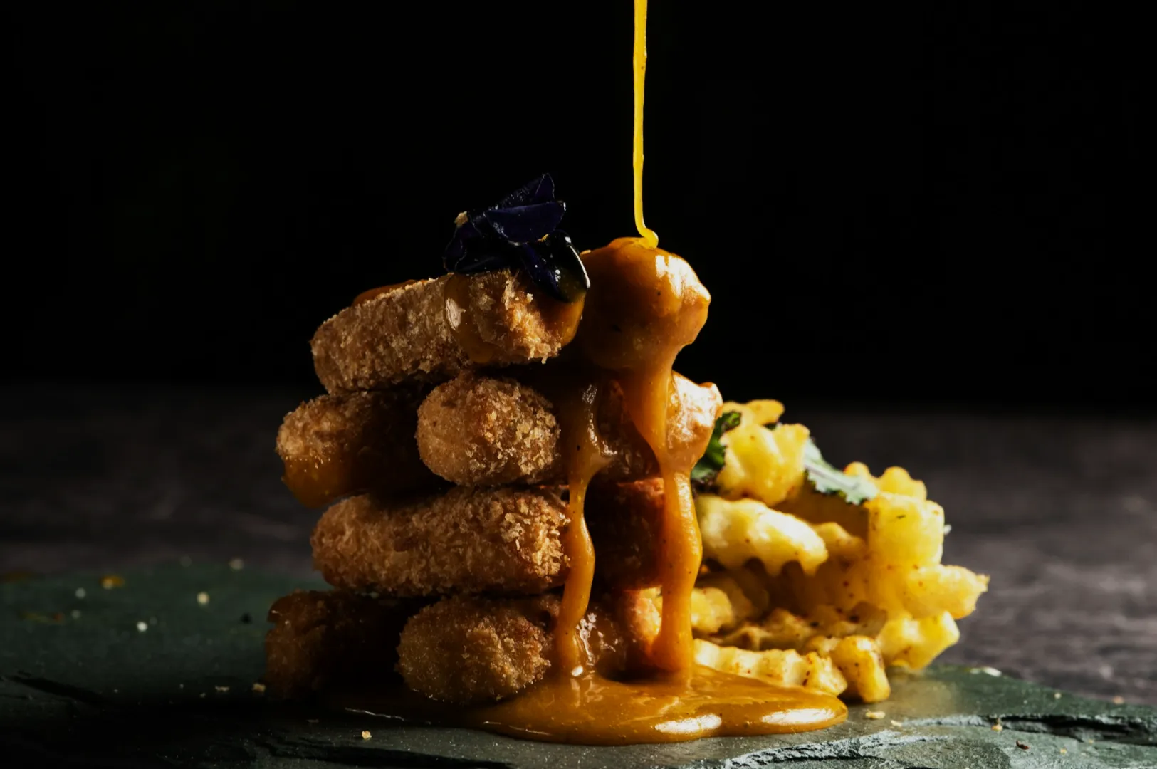Golden sauce drizzles over a stack of crispy croquettes garnished with a flower. Waffle fries are in the background, on a dark plate. Luxurious and appetizing.