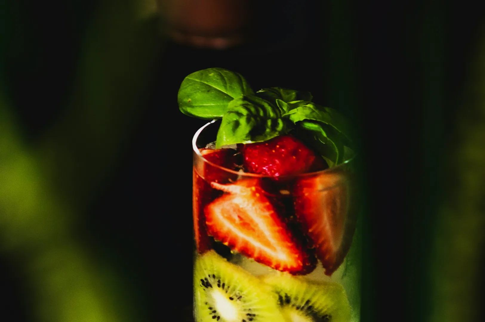 A refreshing drink with sliced strawberries, kiwi, and basil leaves in a clear glass, set against a dark, blurred background.