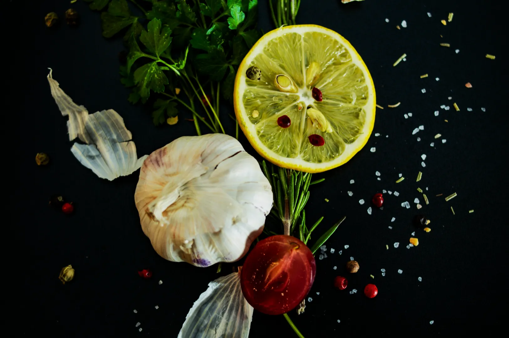 A composition of fresh ingredients on a dark surface, including a lemon slice, garlic, tomato, parsley, rosemary, and scattered spices, suggesting vibrant flavors.