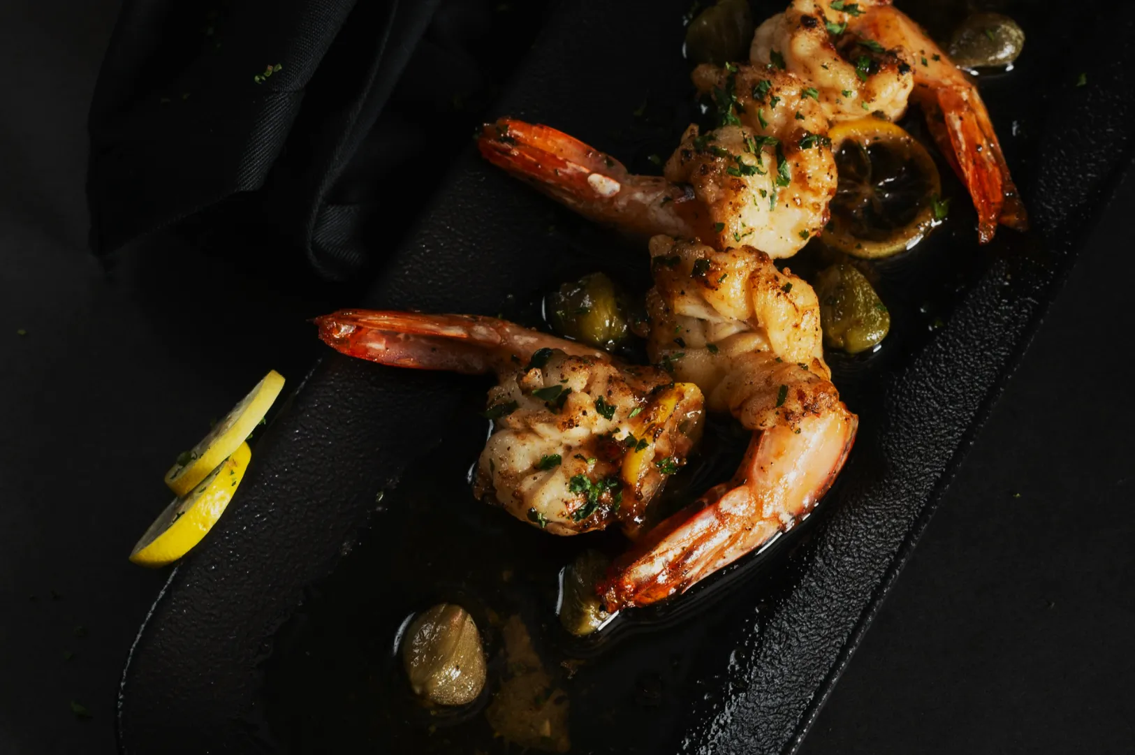 Grilled shrimp garnished with herbs on a black plate, surrounded by lemon slices and capers. The setting is elegant, conveying a gourmet vibe.