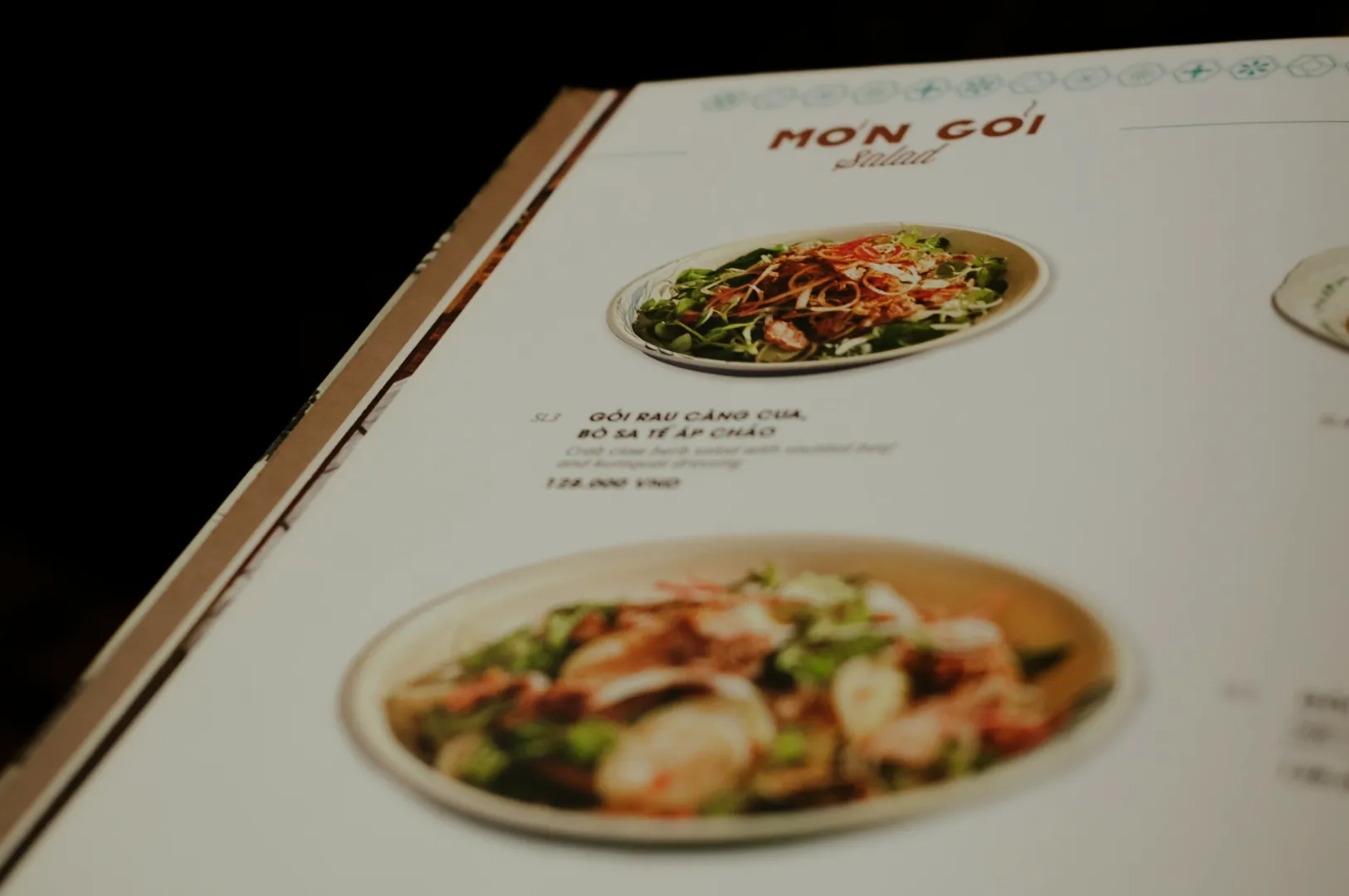 Close-up of a menu with sections labeled "Món Gỏi" and "Salad" featuring two dishes. Each dish is depicted with vibrant greens and garnishes.