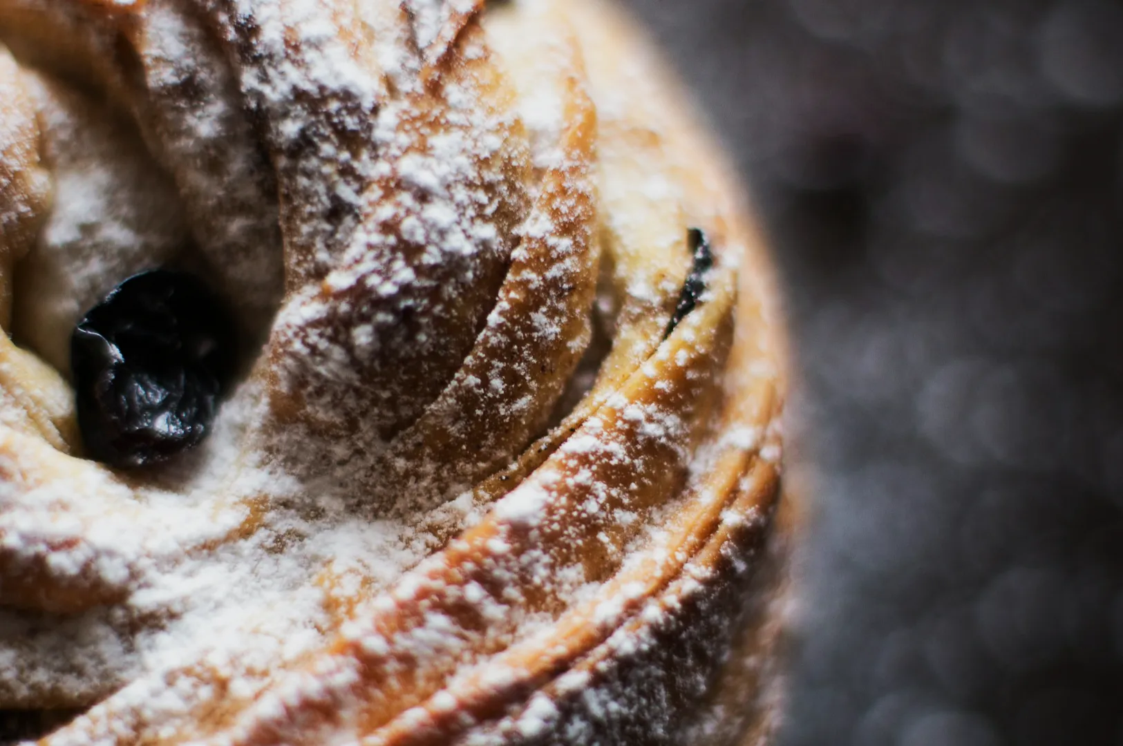 Close-up of a powdered sugar-dusted pastry, highlighting its spiral layers and a visible raisin, set against a blurred, bokeh background.