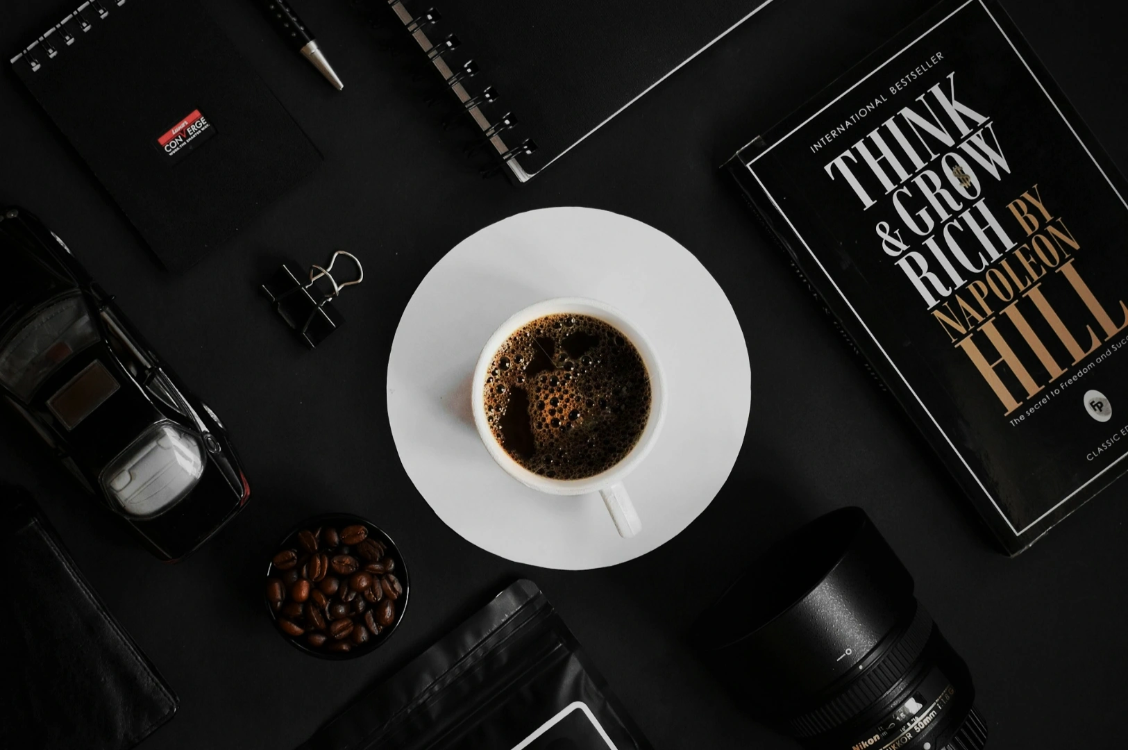 A flat lay of a coffee cup on a saucer surrounded by a book titled "Think & Grow Rich," camera lens, notebooks, a toy car, coffee beans, and a pen on a dark surface. The scene conveys a mood of productivity and creativity.A glass of iced coffee with milk swirls, filled with ice cubes, sits against a dark background. The frothy top adds a refreshing feel.