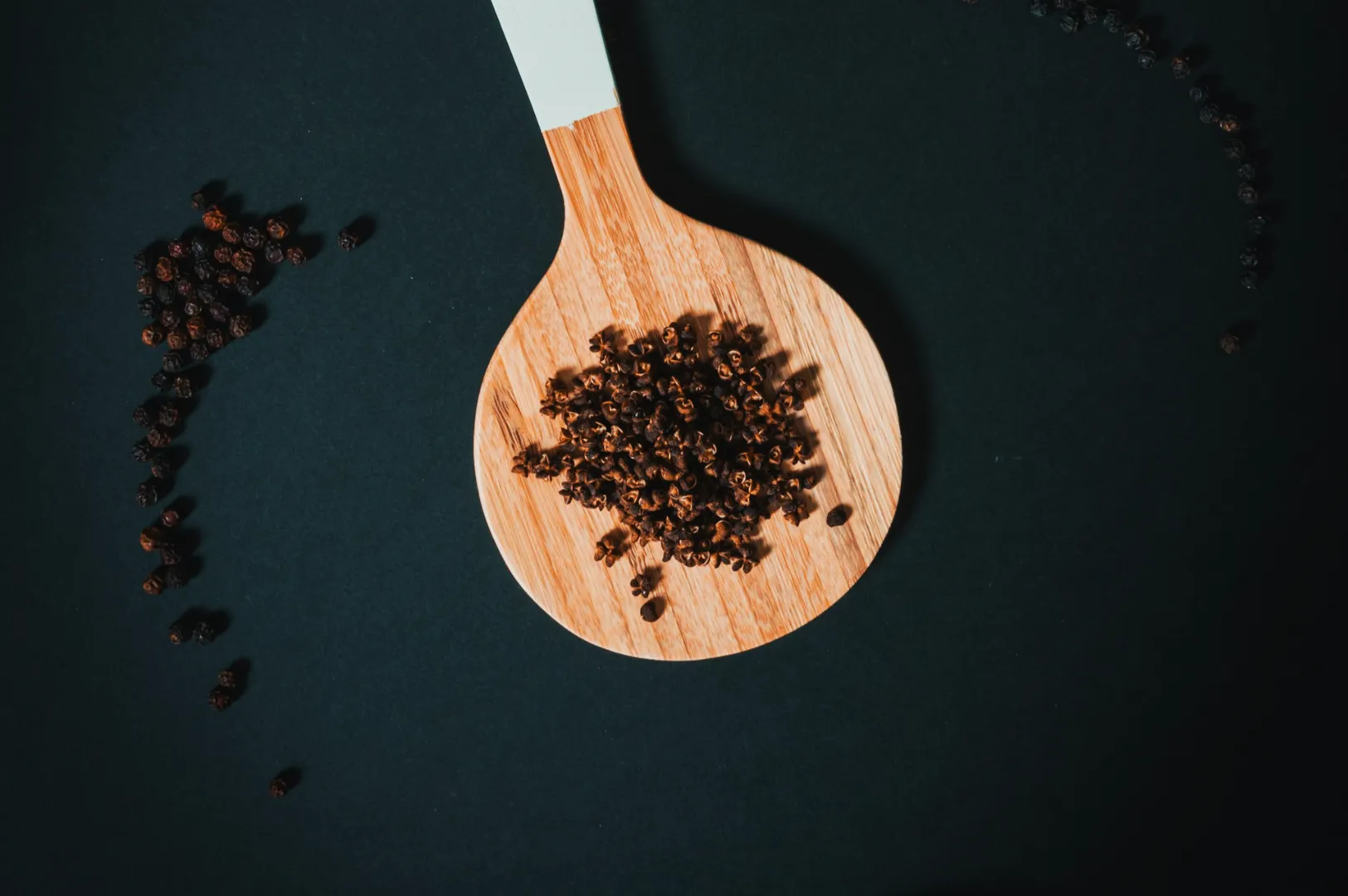 A wooden spoon filled with black peppercorns lies on a dark surface. Scattered peppercorns surround the spoon, creating a rustic and earthy tone.