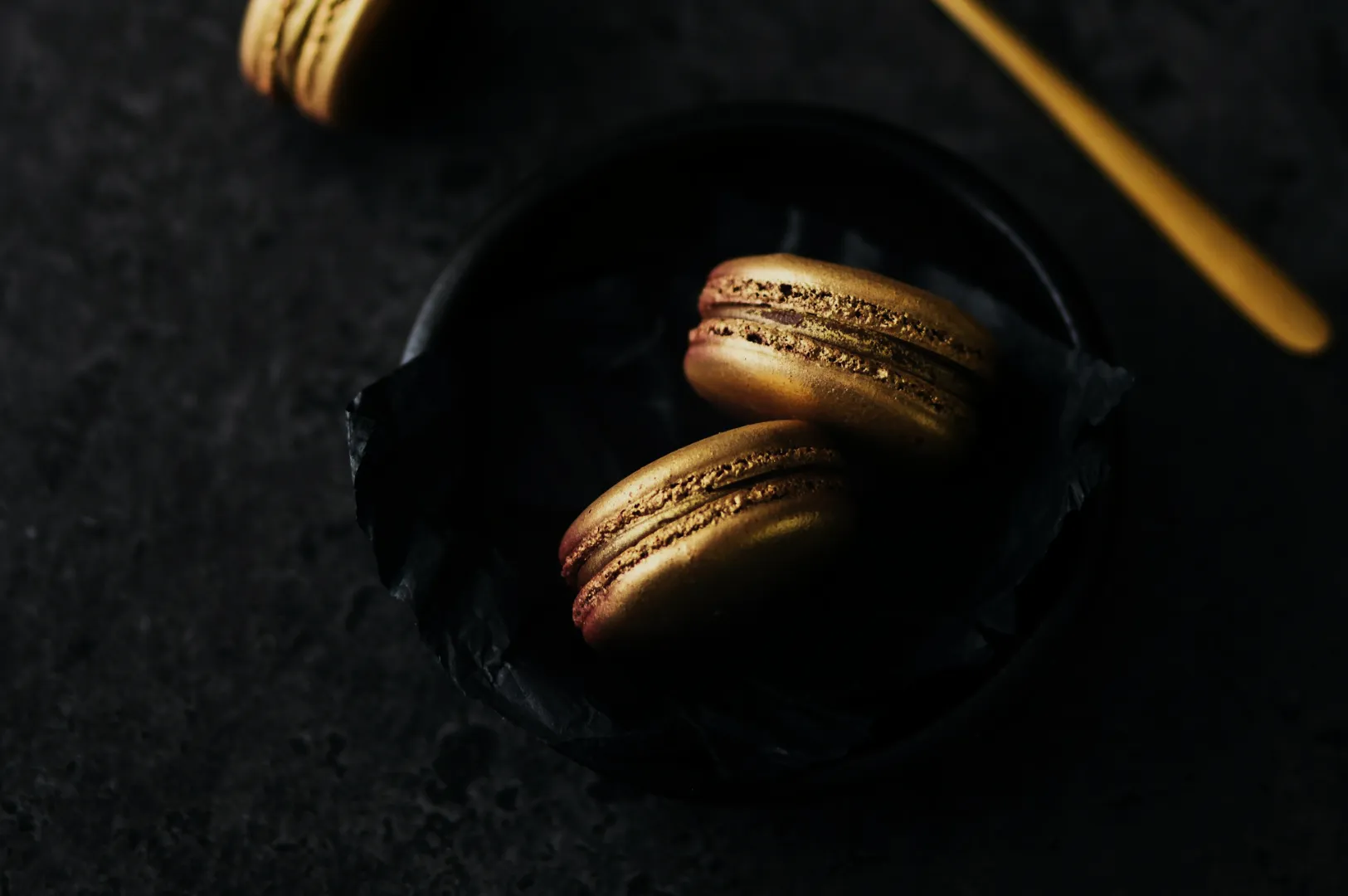 Two golden macarons rest on black paper in a dark bowl. A gold spoon lies beside them on a textured dark surface, creating an elegant, luxurious feel.