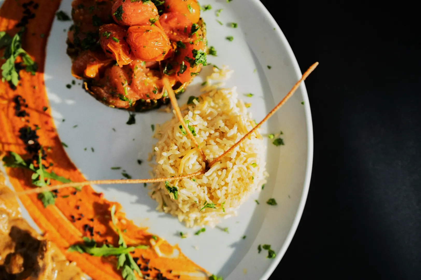 A white plate with a mound of rice garnished with herbs and arranged beside roasted cherry tomatoes. A vibrant, artistic orange sauce swipe decorates the side.