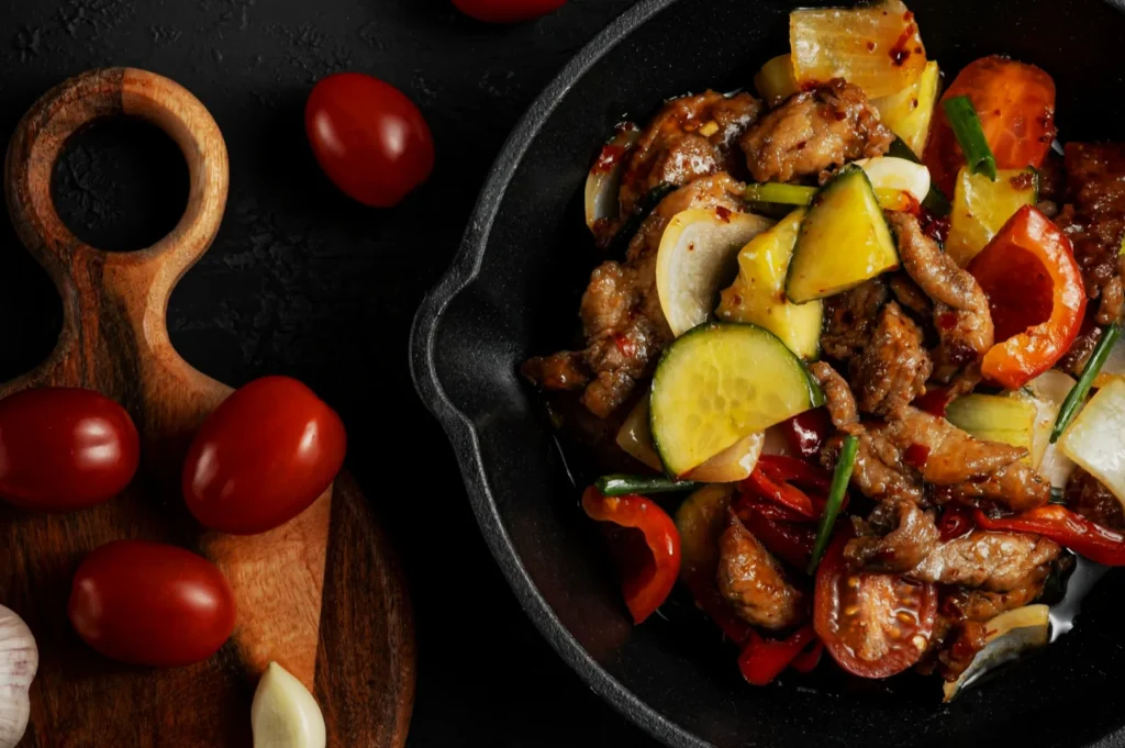 Close-up of a vibrant stir-fry dish in a black bowl, featuring beef, yellow squash, zucchini, onions, and peppers. Nearby, a wooden board holds cherry tomatoes and garlic.