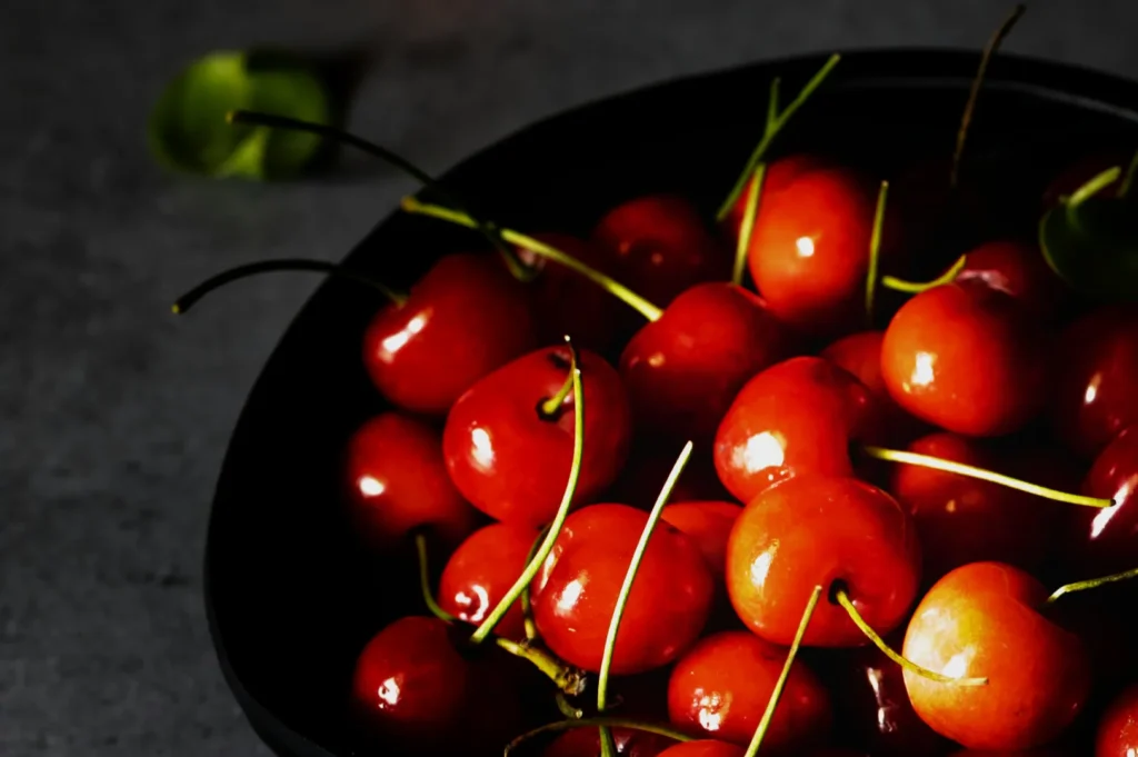 A black bowl filled with glossy red cherries, each with a stem. The moody lighting highlights their rich color and shiny texture against a dark background.