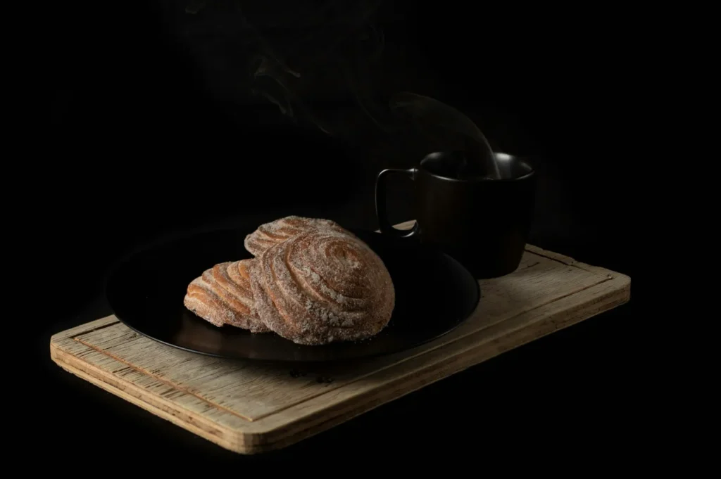 On a wooden board, two sugar-dusted pastries rest on a black plate beside a steaming mug against a dark background, creating a cozy, inviting scene.
