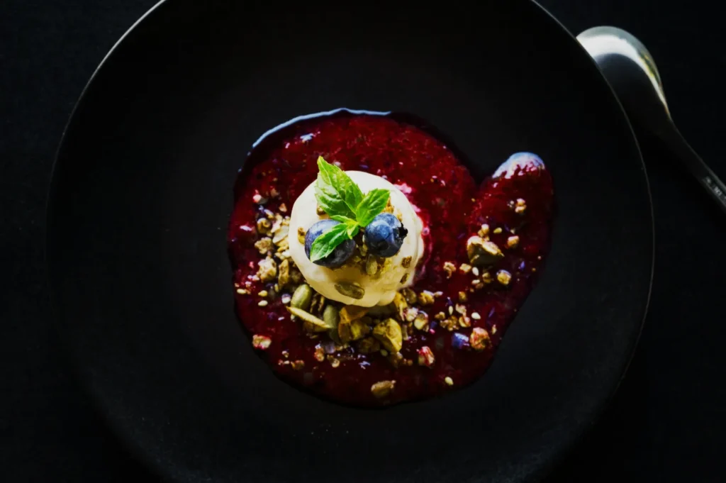 Aerial view of a vanilla ice cream scoop with blueberries and mint on a black plate, surrounded by red berry sauce and crushed nuts, alongside a spoon.
