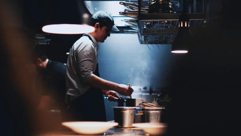 Chef in dimly-lit kitchen focused on cooking, wearing a cap and apron. Steam rises from a pot, creating a warm, bustling atmosphere. Mood is industrious.