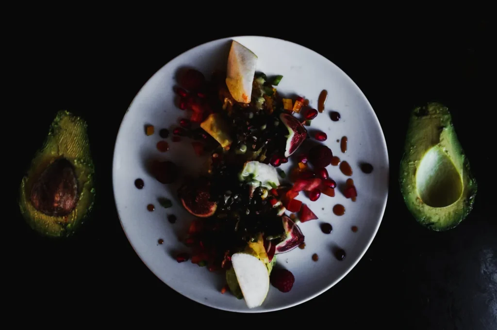 A white plate on a dark table holds a colorful salad with figs, apples, seeds, and pomegranate. Avocado halves are placed beside the plate.