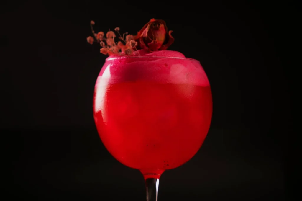 A vibrant red cocktail in a round glass, topped with pink foam and a dried flower garnish, set against a dark background. Elegant and sophisticated.