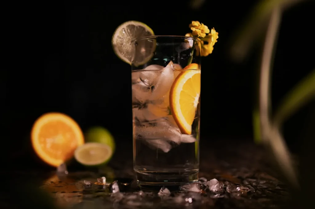 A refreshing drink in a tall glass with ice, orange and lime slices, and a yellow flower garnish. Background has scattered ice and citrus.