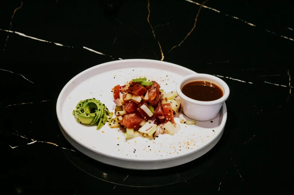 A white plate on a dark marble surface features a colorful poke bowl with diced raw fish, avocado, and vegetables, garnished with black sesame seeds. A small cup of dark sauce sits beside the poke, adding contrast. The arrangement looks fresh and inviting.
