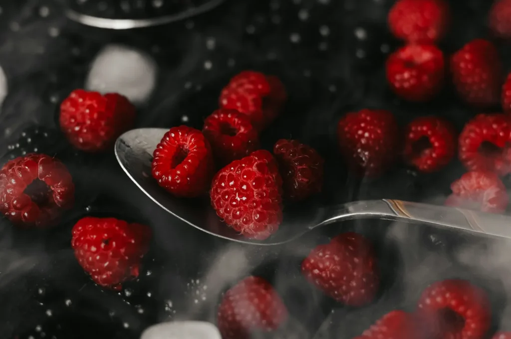 Close-up of fresh, vibrant red raspberries on a silver spoon, surrounded by a misty, smoky ambiance. The dark background enhances their rich color.