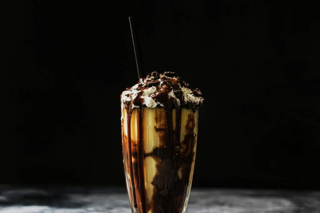 A tall glass of chocolate milkshake topped with whipped cream and chocolate drizzle, set against a dark background. A black straw protrudes from the glass.