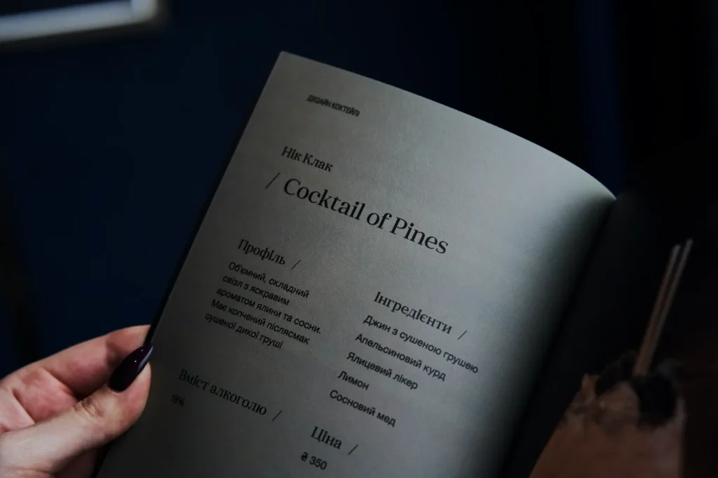 A close-up of a menu page titled "Cocktail of Pines" in both English and another language. It lists ingredients and pricing. A hand with painted nails holds the page.