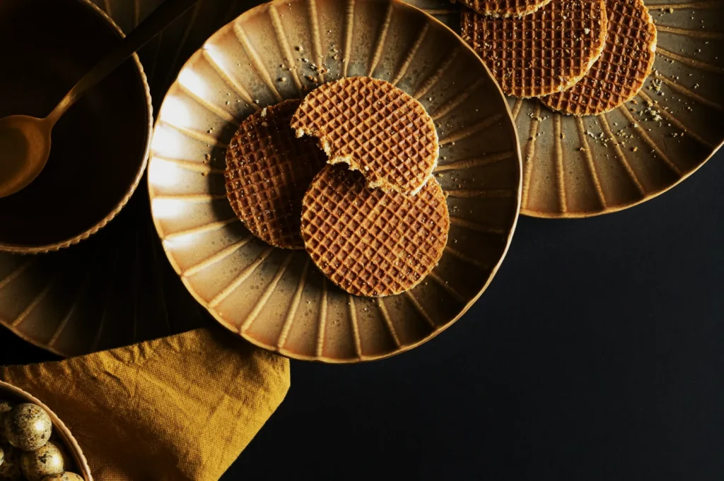 Golden waffle cookies on a textured brown plate with a few crumbs. A contrastful dark background and a mustard cloth add warmth and elegance.