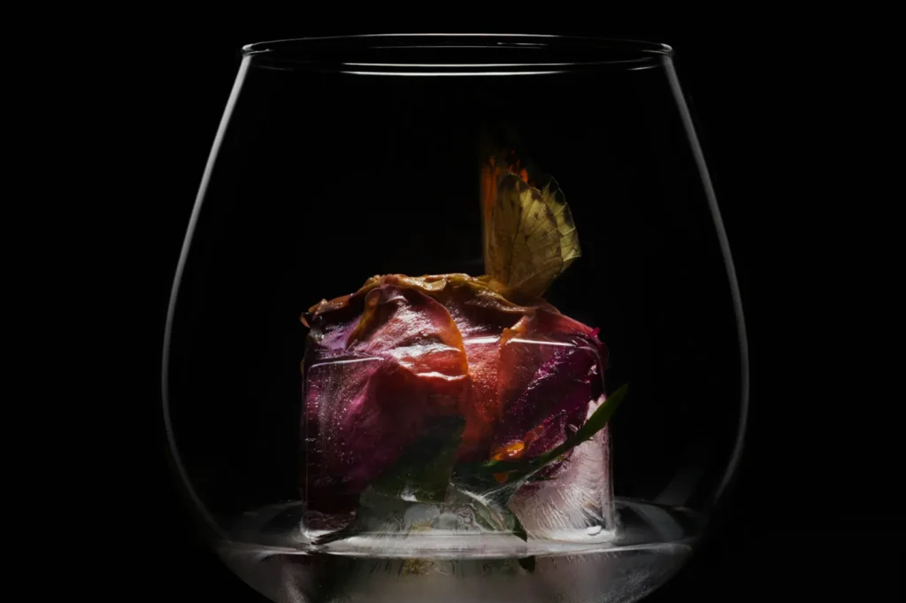 A butterfly and vibrant rose are encased in a clear ice cube, placed inside a large glass. The setting conveys a surreal and elegant mood.