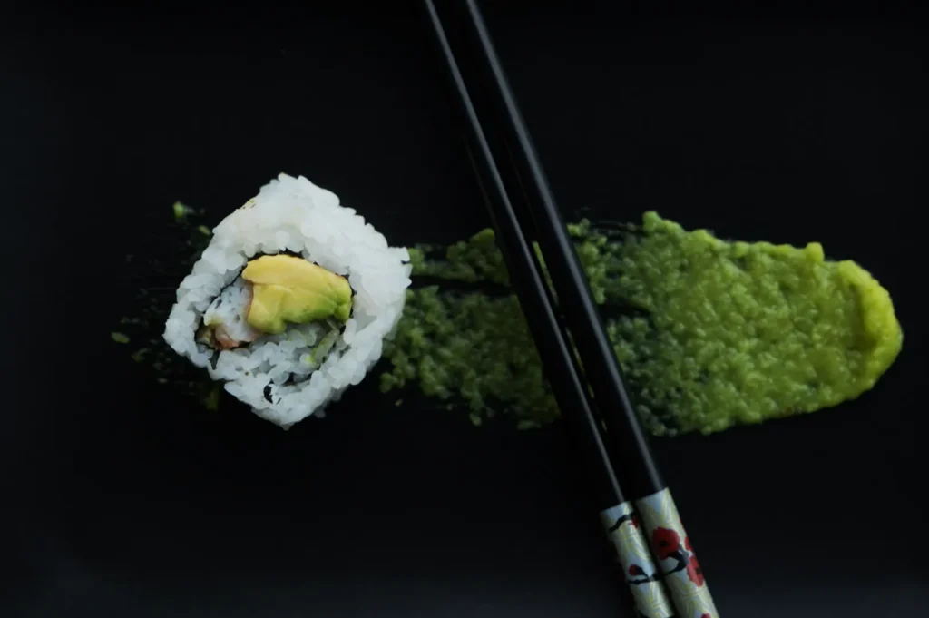 Sushi roll with avocado on black plate, resting on green wasabi spread. A pair of decorated chopsticks lies beside the sushi, creating a minimalist feel.