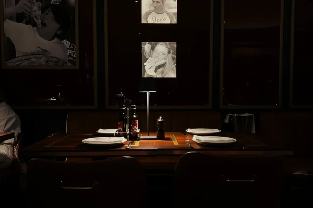 Cozy restaurant booth with dim lighting, set for four. Wall photos of people enjoying food add a lively vibe. Table holds plates, bottles, and a lamp.