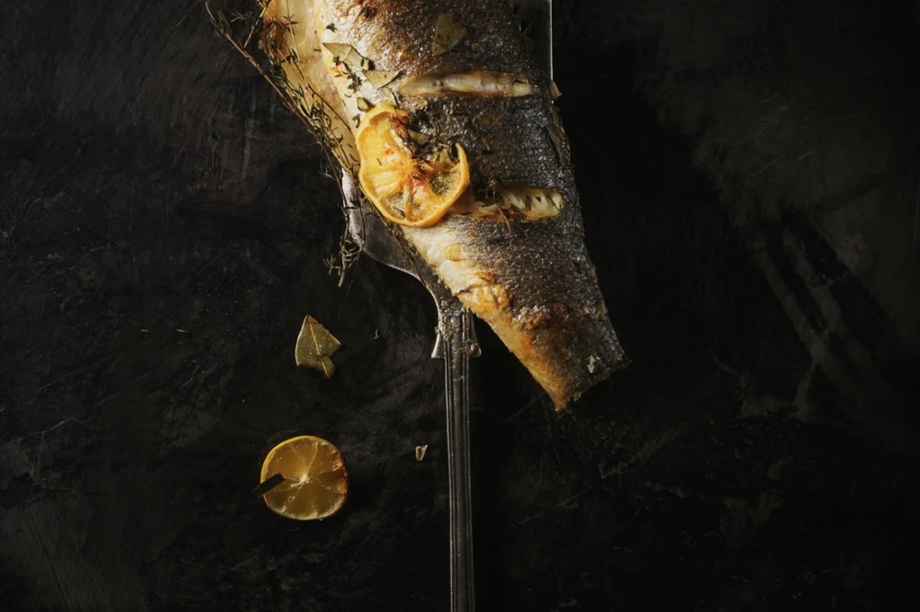Grilled fish garnished with lemon slices and herbs on a rustic dark background. A charred lemon and herb sprigs add a gourmet, rustic feel.