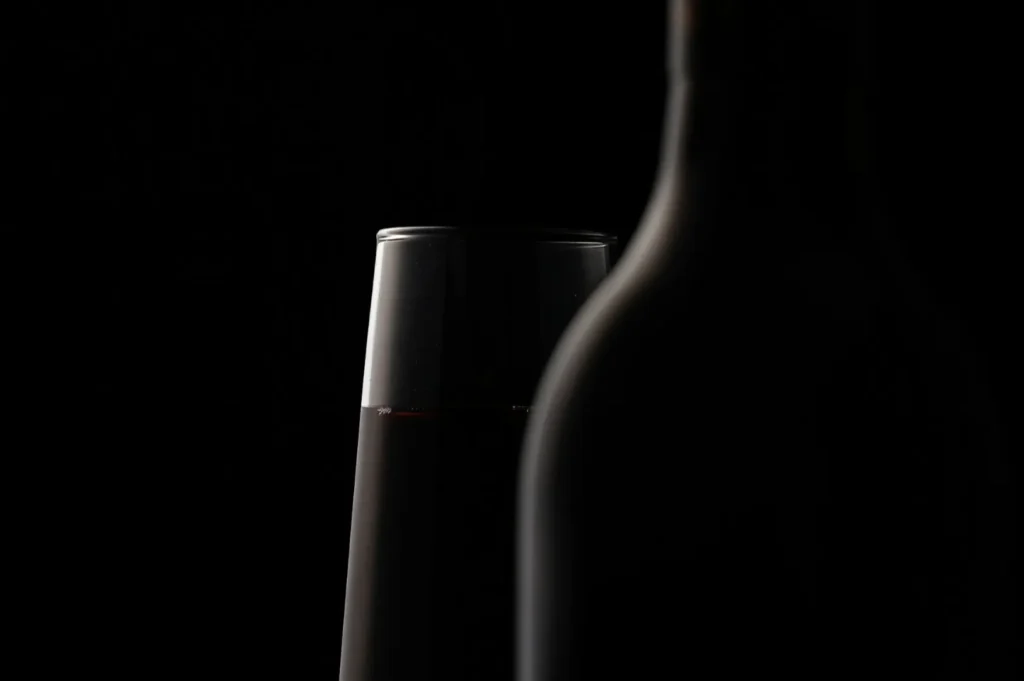 A dimly lit image shows a silhouette of a wine bottle and a glass filled with red wine against a dark background, creating a mysterious, elegant mood.