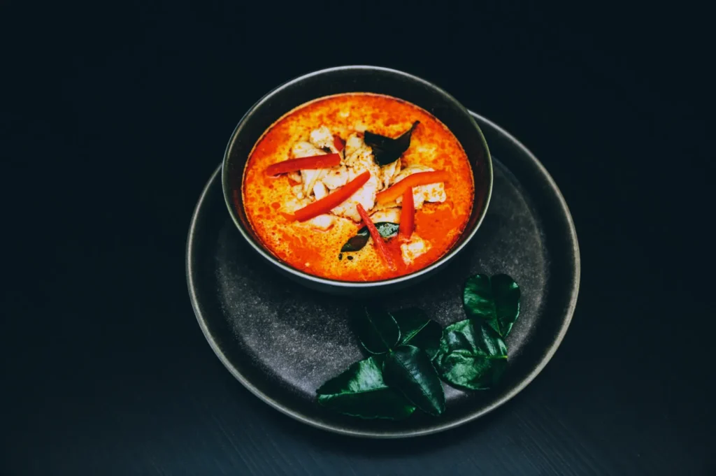 A bowl of vibrant orange Thai curry soup garnished with sliced red peppers and herbs. Displayed on a dark plate with fresh green kaffir lime leaves.