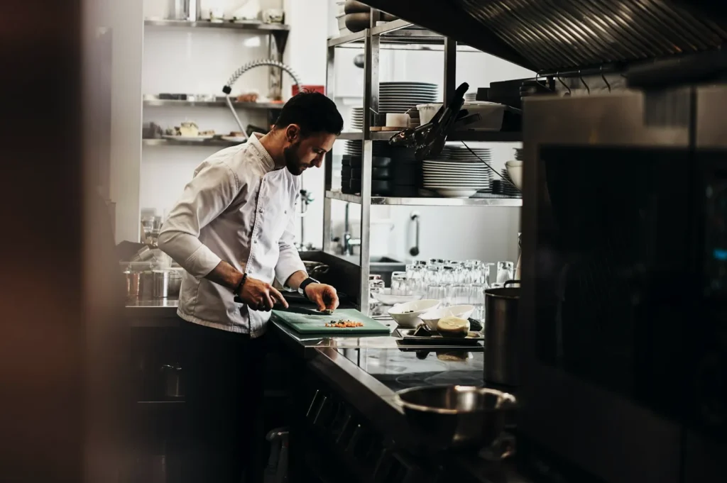 Chef in a professional kitchen cutting vegetables on a green board. Shelves with plates and cutlery are in the background, creating a focused atmosphere.
