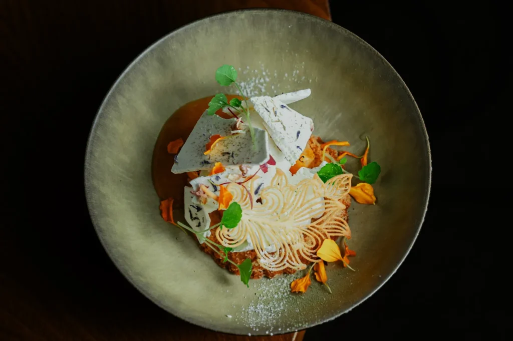 Elegant dessert artfully plated in a bowl with swirls of cream, delicate meringue shards, vibrant orange petals, and green garnishes, evoking a fresh, sophisticated feel.