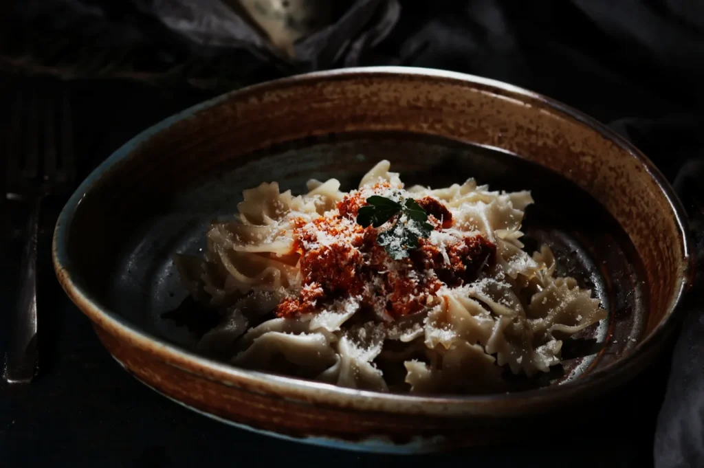 A rustic bowl of farfalle pasta topped with rich tomato sauce, grated cheese, and a basil leaf. The setting conveys a warm, inviting ambiance.