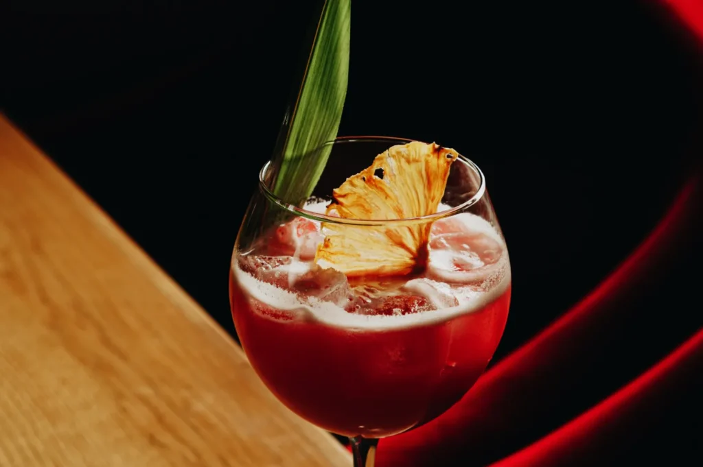 A vibrant red cocktail in a glass, garnished with a pineapple slice and a green leaf, sits on a wooden table against a dark red background.