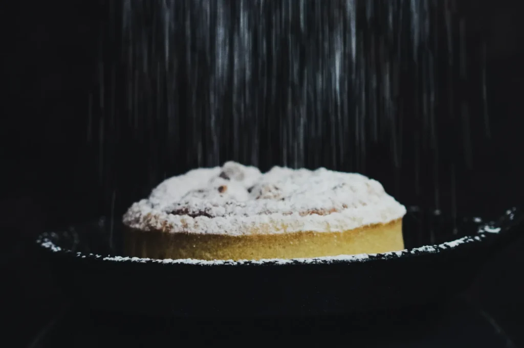 A pie on a dark platter being dusted with powdered sugar, creating a soft, delicate snowfall effect. The mood is cozy and inviting.