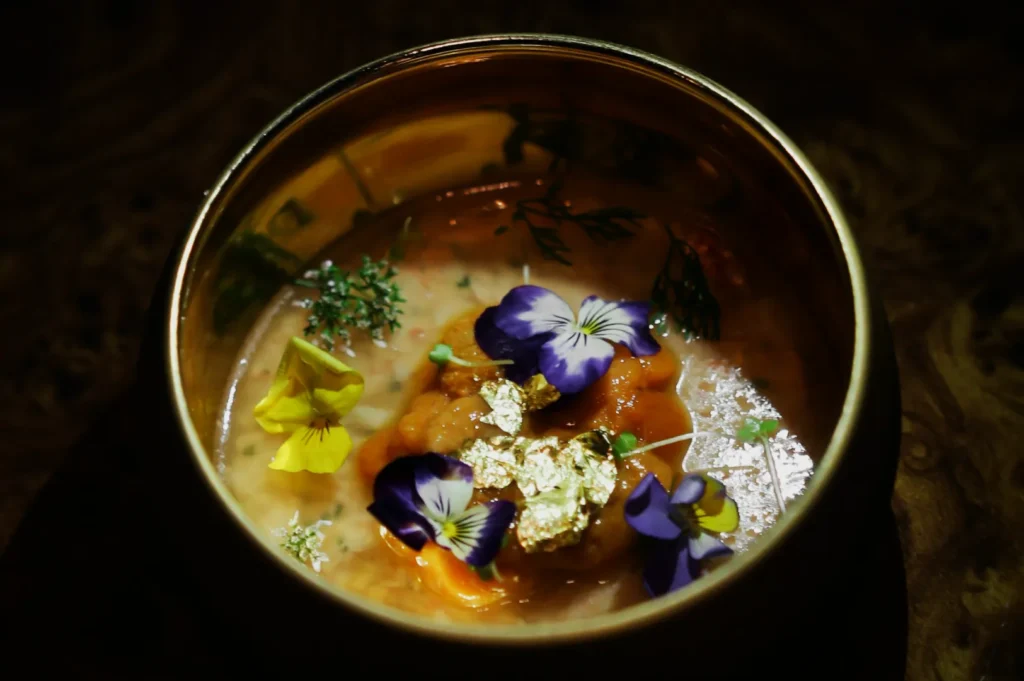 Elegant dish in a round bowl features vibrant purple and yellow edible flowers, garnished with herbs and delicate gold leaf, evoking luxury and artistry.