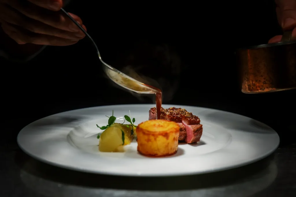 A chef elegantly drizzles sauce over a gourmet dish featuring seared meat, a potato cake, and poached pear, conveying a sense of refinement and artistry.
